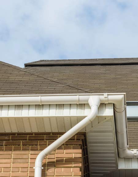 metal-roof Detail of a white gutter and downspout system installed against a brick wall and white siding beneath a brown shingle roof on a home.