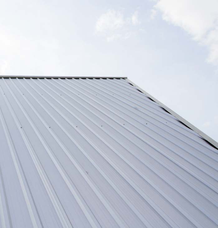 Close-up of a residential or commercial sloped roof installed with new, light-gray, corrugated standing seam metal panels beneath a bright, cloudy sky.