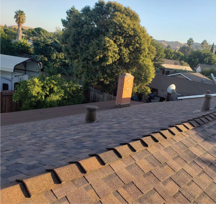 roofers-repairing A warm-toned, multi-layered brown asphalt shingle after roof repair, showing the finished ridge line and surrounding suburban trees and houses.