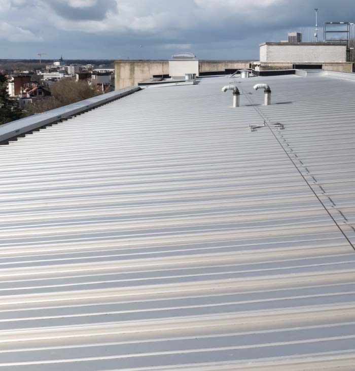 Wide-view of a large, low-slope commercial building roof covered with shiny, corrugated metal roofing, with the city skyline visible in the background.